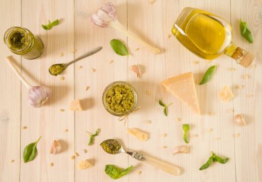 İtalyan fesleğen soslu iki cam kavanoz, pesto soslu iki eski kaşık, parmesan peyniri, fesleğen yaprağı, çam fıstığı, zeytinyağı ve sarımsak beyaz ahşap masada. Üst görünüm