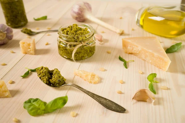 Glass jar full of italian sauce basil pesto and spoon with pesto lay on the white wooden table. Ingredients for traditional pesto: parmesan cheese, basil leaves, pine nuts, olive oil and garlic.