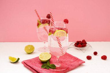 Yaz ferahlatıcı vitamin içecekleri. Limonata ve şeffaf gözlüklü ahududu pembe arka planda beyaz bir masada duruyor. Yanında bir kase ahududu, limon dilimleri ve nane yaprakları var..