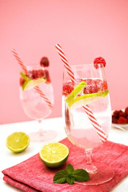 Limonlu ve naneli nefis bir içecek kırmızı tekstil peçetesinin üzerinde ve pembe arka planda beyaz bir masanın üzerinde duruyor. Yanında nane yaprakları ve bir dilim limon var. Bardakta kokteyl tüpü. Dikey