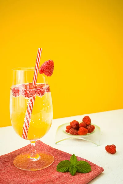 A summer soft drink with raspberries in a glass stands on a red textile napkin and on a white table on an yellow background. Near is a bowl of sweet raspberries and fresh mint leaves. Vertically