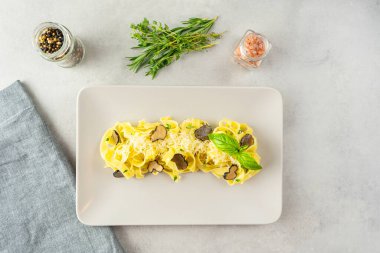 Beyaz tabakta servis edilen siyah trüf mantarlı ve parmesanlı İtalyan Makarnası. Yanında bitkiler (kekik, biberiye), baharatlar (karabiber, pembe mineral tuzu) vardır. Açık gri beton arkaplan, üst görünüm.