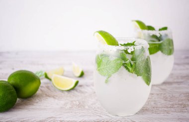 Kireç ve şeffaf bardaklarda naneli ferahlatıcı bir içecek beyaz bir duvarın arka planında, ahşap bir masanın üzerinde duruyor. Hemen yanında sulu limon dilimleri ve çizgili kokteyl tüpleri var..