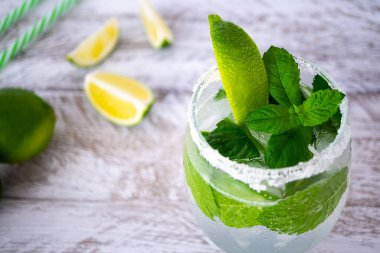 Şeffaf bardakta mojito kokteyli, buz, nane yaprakları ve açık ahşap zemin üzerinde bir dilim limon. Bardağın yanında üç dilim sulu limon var. Bardağın kenarları tuzla kaplıdır, yakın plan..