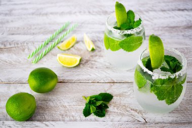 Yaz ferahlatıcı limonlu içki ve güzel bardaklarda nane şekeri açık ahşap bir masada duruyor. Yakınlarda iki limon, nane yaprakları ve kokteyl tüpleri var. Bardağın kenarları tuzla kaplıdır..