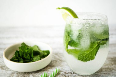 Kireç ve nane arkaplanlı, ahşap arka plan, klasik ve pastel renkte bir bardakta ev yapımı kokteyl. Yanında naneli bir kase var. Detoks sabah içeceği, temiz yemek..