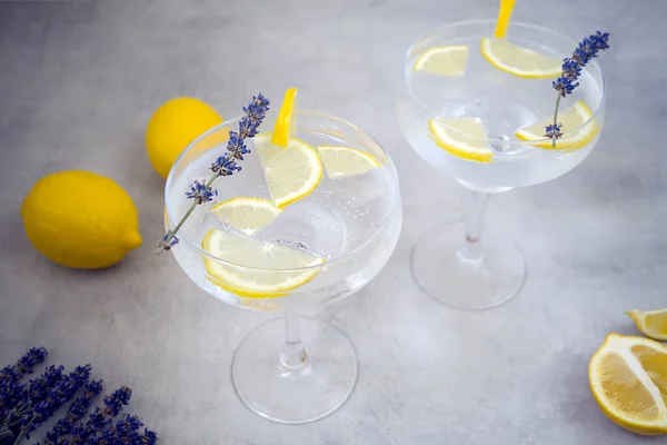 Lemon non-alcoholic cocktails with ice cubes in beautiful glasses for margaret stands on a gray concrete background. Glasses are decorated with lavender and a slice of lemon. Nearby lies two lemons.
