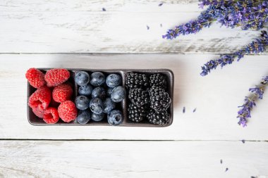Olgun böğürtlen (ahududu, böğürtlen, yabanmersini) farklı dallarda siyah bir tabakta bulunur. Beyaz ahşap bir arka planda duran bir tabak. Sağ üst köşede lavanta çiçekleri var. Üst görünüm