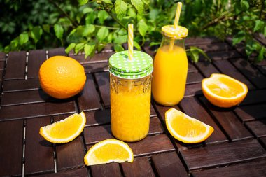 Cam bir kavanozda taze sıkılmış portakal suyu ve açık havada kahverengi bir ahşap bahçe masasında duran bir şişe yeşil yaprakların arka planında duruyor. Sulu portakal dilimleri yakınlarda duruyor. Sağlıklı beslenme, vitaminler.