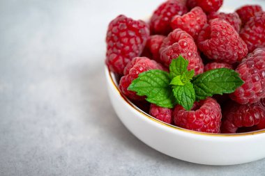 Beyaz bir kasede, üzerinde altın bir kenarı olan nane yaprakları olan olgun ahududular gri bir arka planda duruyor. Sağlıklı tatlı, düzgün beslenme. Mevsimlik taze meyveler. Yakın plan.