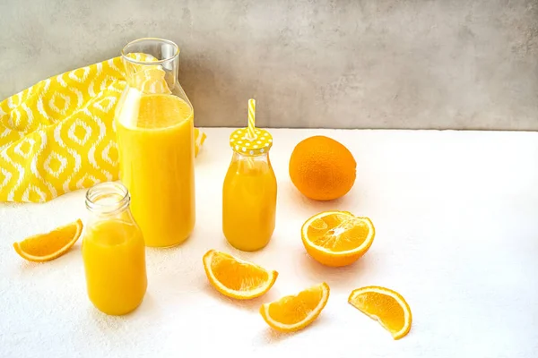新挤压的健康橙汁在瓶子和酒瓶中 白色的桌子上靠着灰色的混凝土墙 旁边躺着多汁的橙子片和一条带白色图案的黄色毛巾 — 图库照片