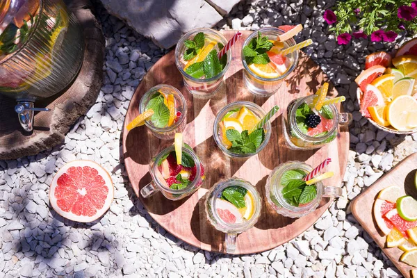 Bright summer cocktails with fruits (grapefruit, lime, lemon) and mint in glass glasses stand on a round wooden tray on the stones in the garden. Summer day, sun rays and shadows. Top view