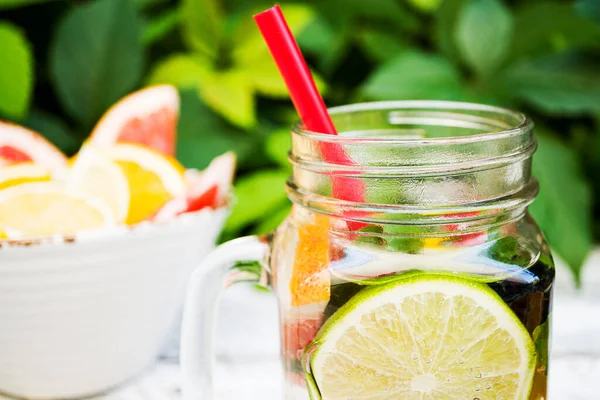Glass cocktail mug with lemonade with slices of grapefruit, lime and lemon stands on a background of green foliage in an outdoor garden. In the mug is a red cocktail straw. Close-up