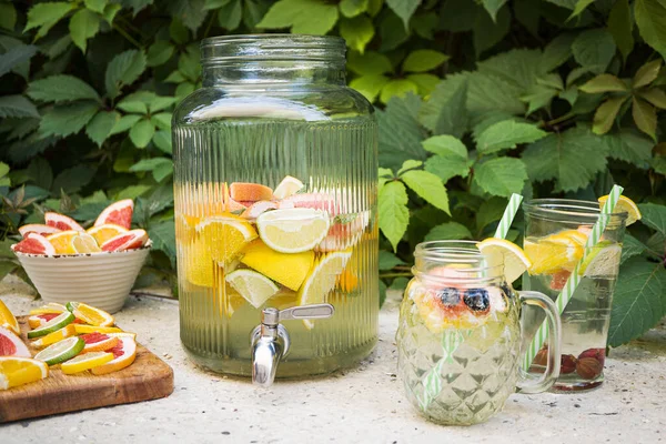 A large jug with a drink and fruit slices (lemon, lime, grapefruit) stands in the garden against a background of green foliage. Nearby are a mug and a glass with a drink, a bowl with slices of fruit.