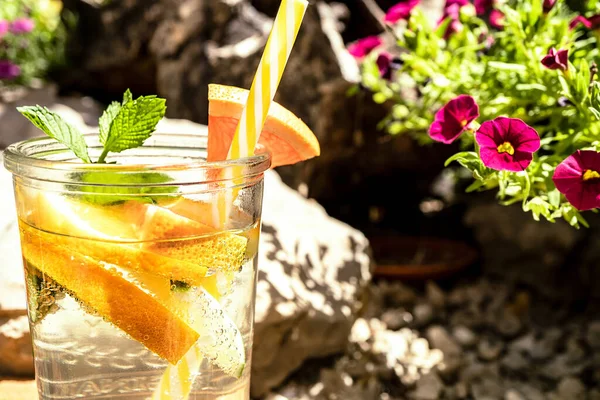 A refreshing champagne-based cocktail with fruits (orange, lime) and mint in a glass is standing outdoors in the garden. The glass is decorated with an orange slice and a cocktail straw. Close-up