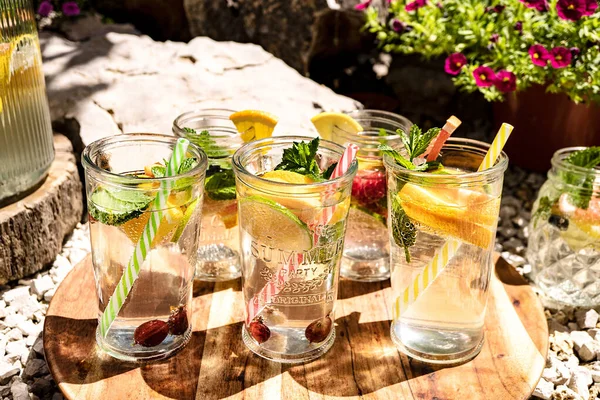 Refreshing Infused water with fruits (lemon, lime, orange), rose hips and mint in glasses stands on a wooden round tray in the open air in the garden. Detox diet, vitamin smoothies, proper nutrition