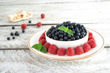 Beyaz bir kase sulu yaban mersini beyaz bir uzay aracının üzerinde, açık ahşap bir masada altın bir kenarı var. Tezgah tabağı olgun ahududu ve nane yapraklarıyla süslenmiş. Mevsimlik böğürtlen, güzel sunum