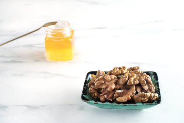 Siyah desenli yeşil bir kasede soyulmuş ceviz, beyaz bir masada duruyor. Yakınlarda küçük cam bir kavanozda bal var. Doğal organik ürünler. Günde bir avuç fındık yemek çeşitli hastalıkları önleyebilir.