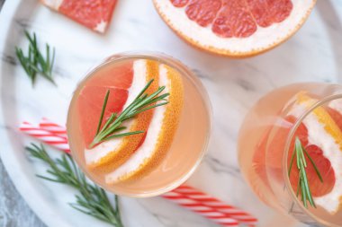 İki bardak ferahlatıcı içecek, greyfurt dilimleri ve biberiye filizleri beyaz tepside duruyor. Yakınlarda çizgili kokteyl tüpleri var. Yaz içkilerini tazeliyorum. Üst görünüm, yakın çekim