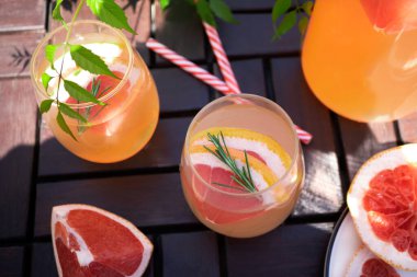 Bahçedeki kahverengi ahşap bir masada, greyfurtlu ve biberiyeli yaz kokteyli duruyor. Yakınlarda greyfurt kamaları ve çizgili kokteyl çubukları var. Güneş ve gölgeler. Üst görünüm