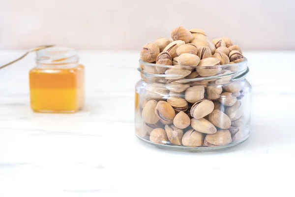 Fried salted pistachios in a glass transparent jar stand on a white surface. In the background is a small jar of honey. Natural organic products concept. Delicious snacks