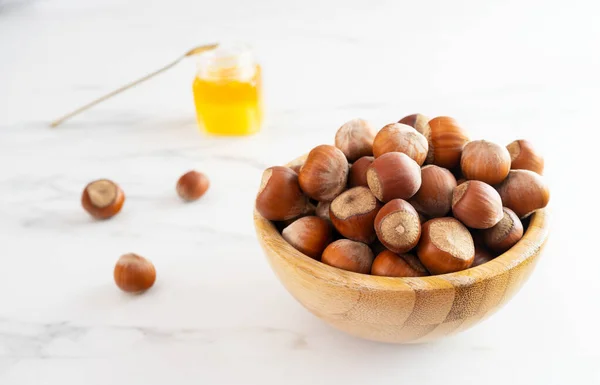 Hazelnuts in a shell in a beautiful wooden bowl stands on a white table. In the background is a small jar of honey and a few nuts. Healthy food, natural organic products. Close-up, selective focus