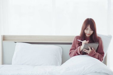 Beyaz bir yatak odasında tablet çalan bir kadın.