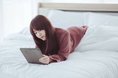 Beyaz bir yatak odasında tablet çalan bir kadın.