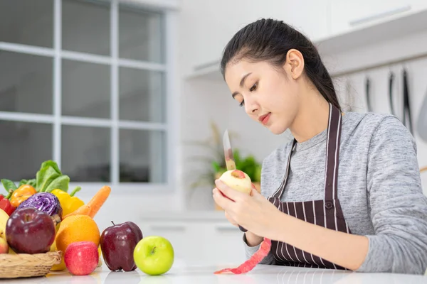 Beautiful asian woman, housewife peeling red apple in white kitchen with plenty of fresh vegetables and fruits on the table.
