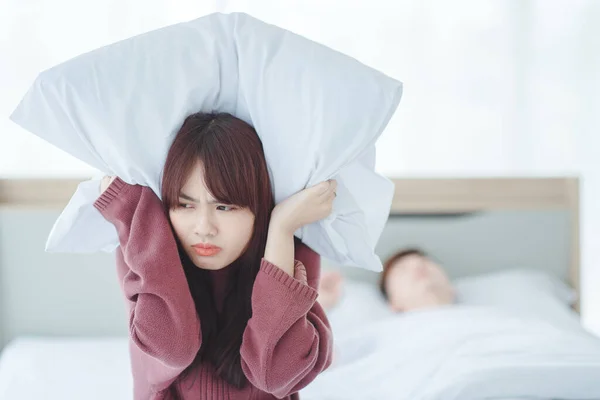 couple in bed man sleeps and snores while woman is unable to sleep closing her ears with big pillows,woman blocking ears with pillow while man snoring on bed