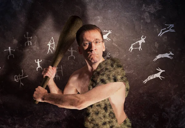 A modern man wearing glasses in primitive clothes with a cudgel in his hand on the background of a stone wall with rock paintings. The metaphor of political threats of the modern world