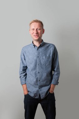 Gri gömlekli neşeli bir adam ayakta ve gülümseyerek. Elleri ceplerinde. İzole