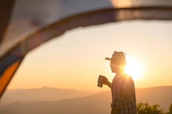 レジャーと自由を楽しんでいる夕日の光空の下山地周辺テント近くでコーヒー カップ滞在を保持するいると幸せな男 — ストック写真