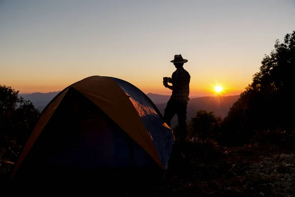 レジャーと自由を楽しんでいる夕日の光空の下山地周辺テント近くでコーヒー カップ滞在を保持するいると幸せな男のシルエット — ストック写真