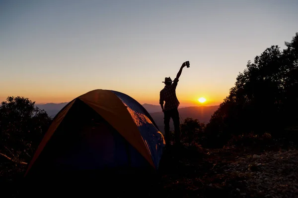 レジャーと自由を楽しんでいる夕日の光空の下山地周辺テント近くでコーヒー カップ滞在を保持するいると幸せな男のシルエット — ストック写真