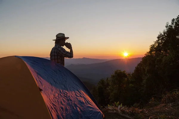 レジャーと自由を楽しんでいる夕日の光空の下山地周辺テント近く飲むと幸せな男 — ストック写真