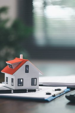 Emlakçı ve müşteri ev satın alma sözleşmesini tartışırken, arka planda emlak, sigorta ve kredi düzenlemelerini sembolize eden ev modeli süreçte yer alıyor.