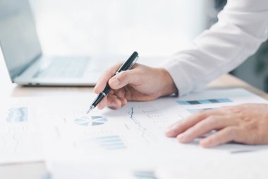 İşadamı mali raporları gözden geçirir, çizelgede kalemle işaret eder, ofis masasındaki verileri analiz eder ve doğruluk için hesap makinesiyle çalışır. Muhasebe kavramı, iş planlaması, yatırım stratejisi.