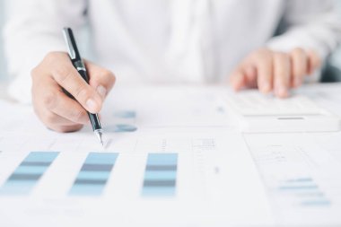 İşadamı mali raporları gözden geçirir, çizelgede kalemle işaret eder, ofis masasındaki verileri analiz eder ve doğruluk için hesap makinesiyle çalışır. Muhasebe kavramı, iş planlaması, yatırım stratejisi.