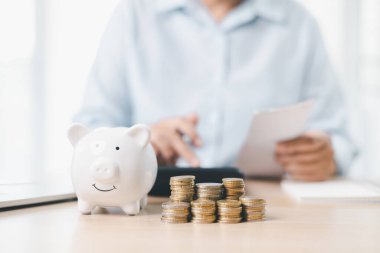 Tasarruf ve yatırım planlama kavramı. Bir iş kadını, mali güvenliği, bütçe yönetimini ve gelecekteki zenginliği sembolize eden belgelerle birlikte ofis masasındaki beyaz domuz kumbarasına para atıyor..