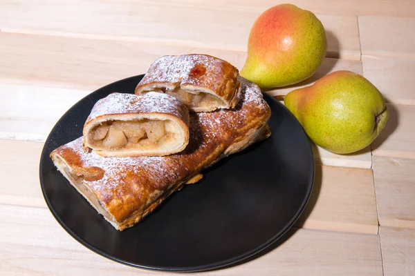 Strudel with apples on a black and white plate with yellow pears on a wooden board, closeup