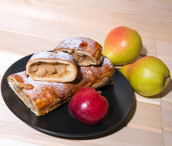 Strudel with apples on a black and white plate of yellow-red peaches, pears on a wooden board, closeup