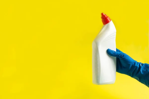 Hand in a rubber blue glove holds sponge and plastic bottle of liquid detergent on a yellow background. Concept of cleaning the house, dusting, housework. Cleaning business, production