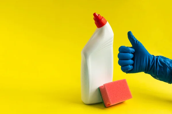 Hand in a rubber blue glove holds sponge and plastic bottle of liquid detergent on a yellow background. Concept of cleaning the house, dusting, housework. Cleaning business, production