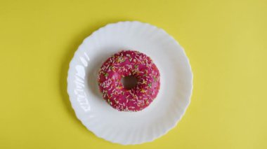 Bir kadının eli, üzerine renkli şekerlemeler serpiştirilmiş pembe bir donuta uzanır ve parlak sarı arka planda beyaz bir tabaktan çörek almaya karar verir. Sağlıksız yiyecekler. Üst görünüm.