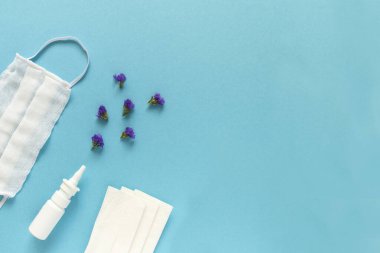 Mevsimlik bahar alerjisi çiçek konsepti. Beyaz sprey kutusu, mor çiçekli peçeteler ve mavi arka planda tıbbi koruyucu maske. Yaratıcı düz konum, kopyalama alanı, asgari biçim