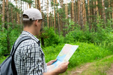 Gri-beyaz tişört şapkalı, gri sırt çantalı, ormanda bir patika boyunca yürüyen, mesafeye bakan bir erkek turistin arka görüntüsü. Elinde harita olan bir turist. Tatil ve doğa aktiviteleri kavramı.