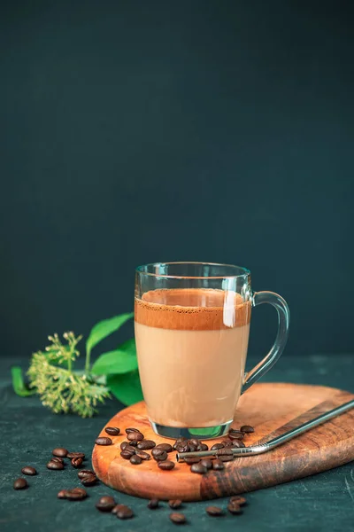 Café Dalgona con espuma en taza de vidrio sobre tabla de cortar, granos de café y paja de metal, fondo negro —  Fotos de Stock