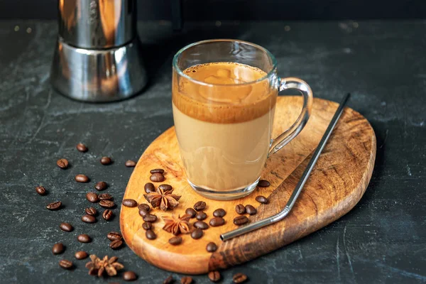 Café Dalgona avec mousse dans une tasse en verre sur planche à découper, grains de café et paille métallique, fond noir — Photo