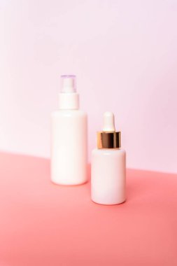 Beyaz duvara karşı pembe masada iki şişe serum ve kozmetik losyon. Damgasız boş paketler. Doğal cilt bakım ürünleri konsepti. Seçici odak